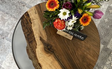 Ein runder Tisch aus dunklem Holz und Epoxidharz. Darauf steht ein üppiger Strauß Blumen und ein Schild, das halb verdeckt ist. Auf einem Schild kann man "brocante" lesen.
