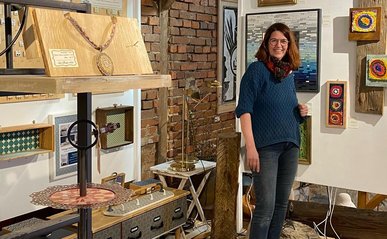 Eine Frau, Andrea Hiller, steht in ihrem Atelier umgeben von Mosaikkunst und lächelt in die Kamera.